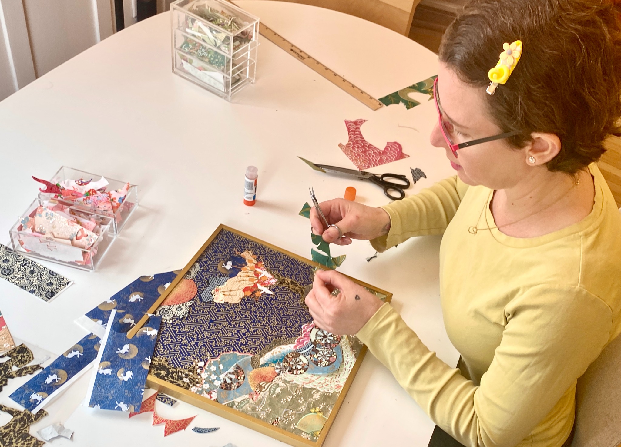 Reut working from above at a white studio table, cutting a small green piece of patterned paper with scissors among paper fragments and an artwork in progress