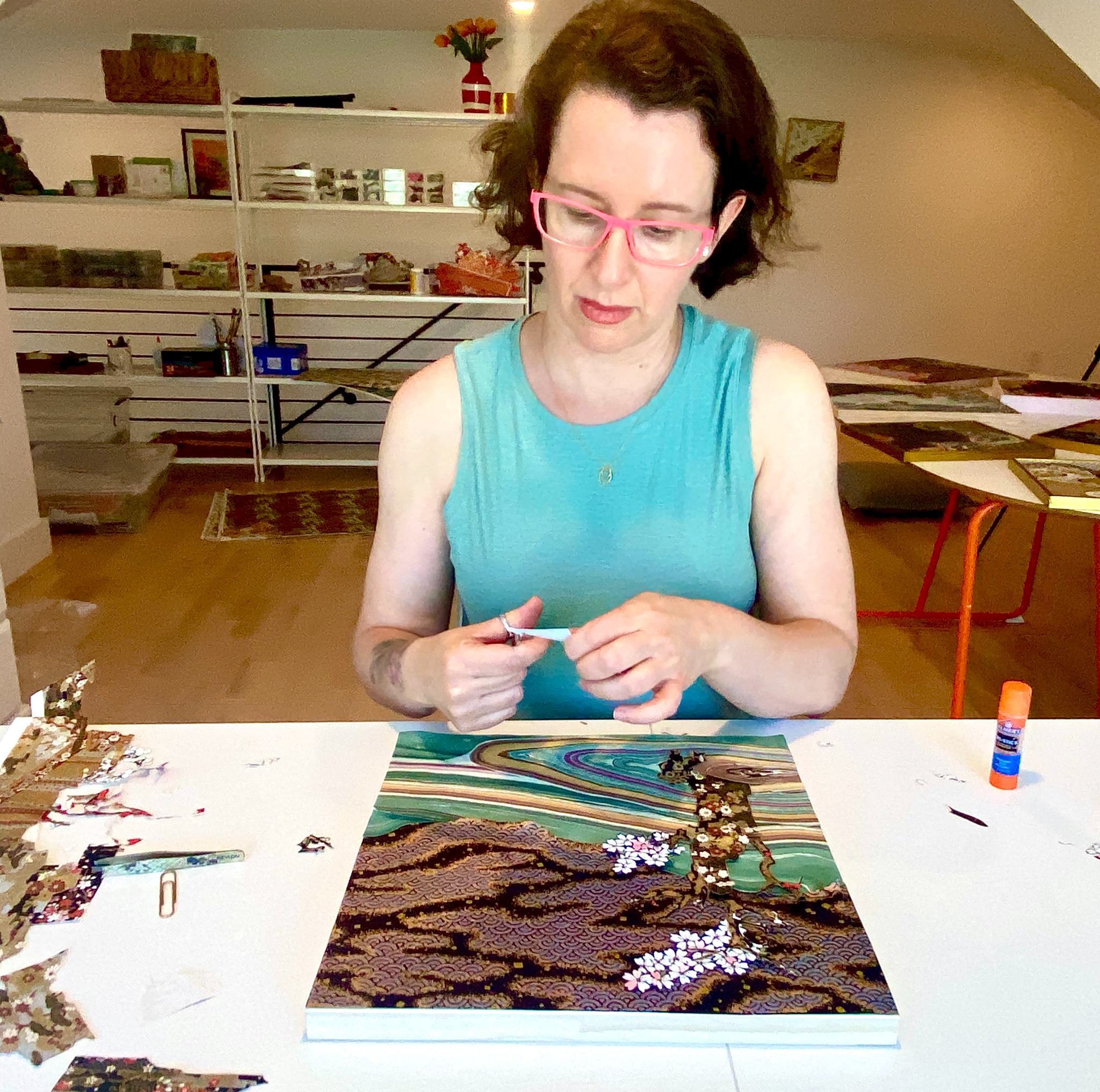 Reut Akerman working at a white studio table, cutting patterned paper with scissors beside an artwork in progress and surrounding materials